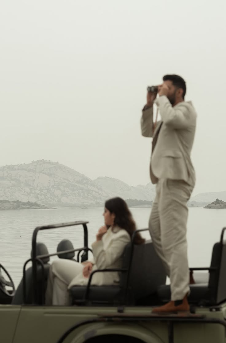 Beige suit with binoculars beside a seated partner on a jeep at the lake