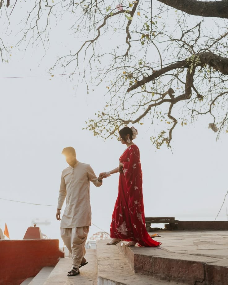 Red floral saree and neutral kurta walking under bare trees by the ghats